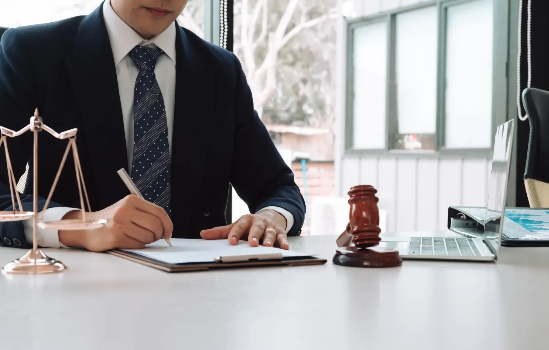 Professional lawyer in formal business attire writing on documents at desk with scales of justice and gavel, symbolizing lega